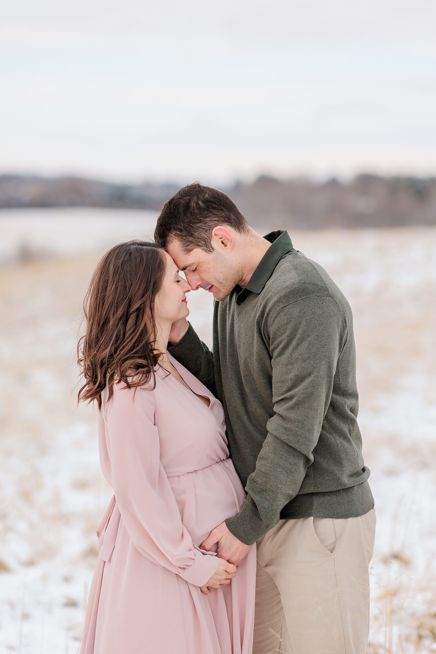 couple stands together touching foreheads and holding hands during snowy maternity session