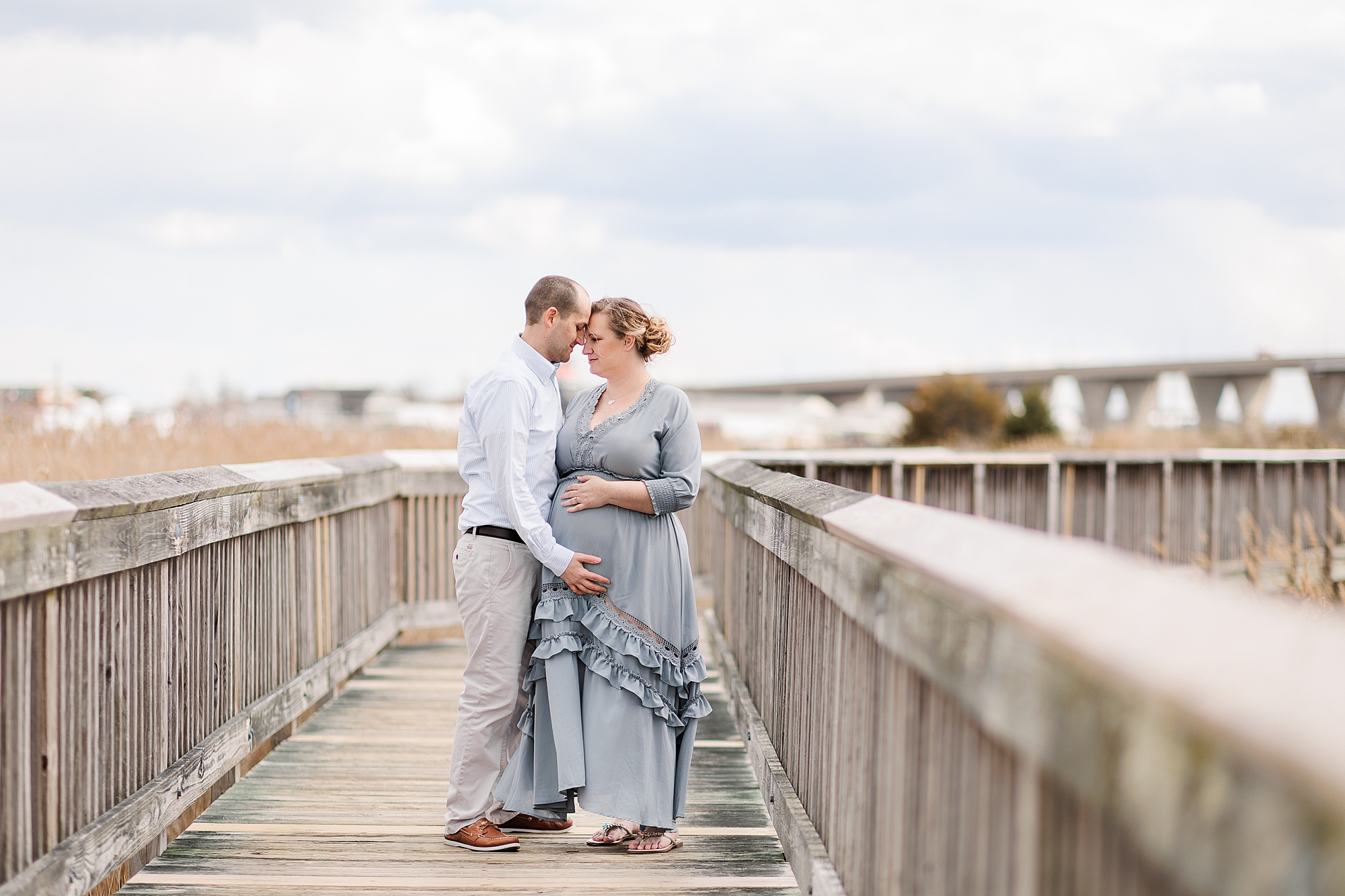 husband and wife stand on wooden pier with mom in ruffled blue dress