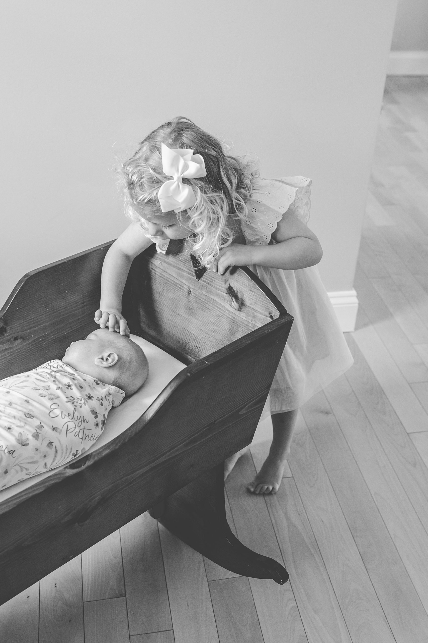 big sister leans over edge of wooden cradle touching baby's head