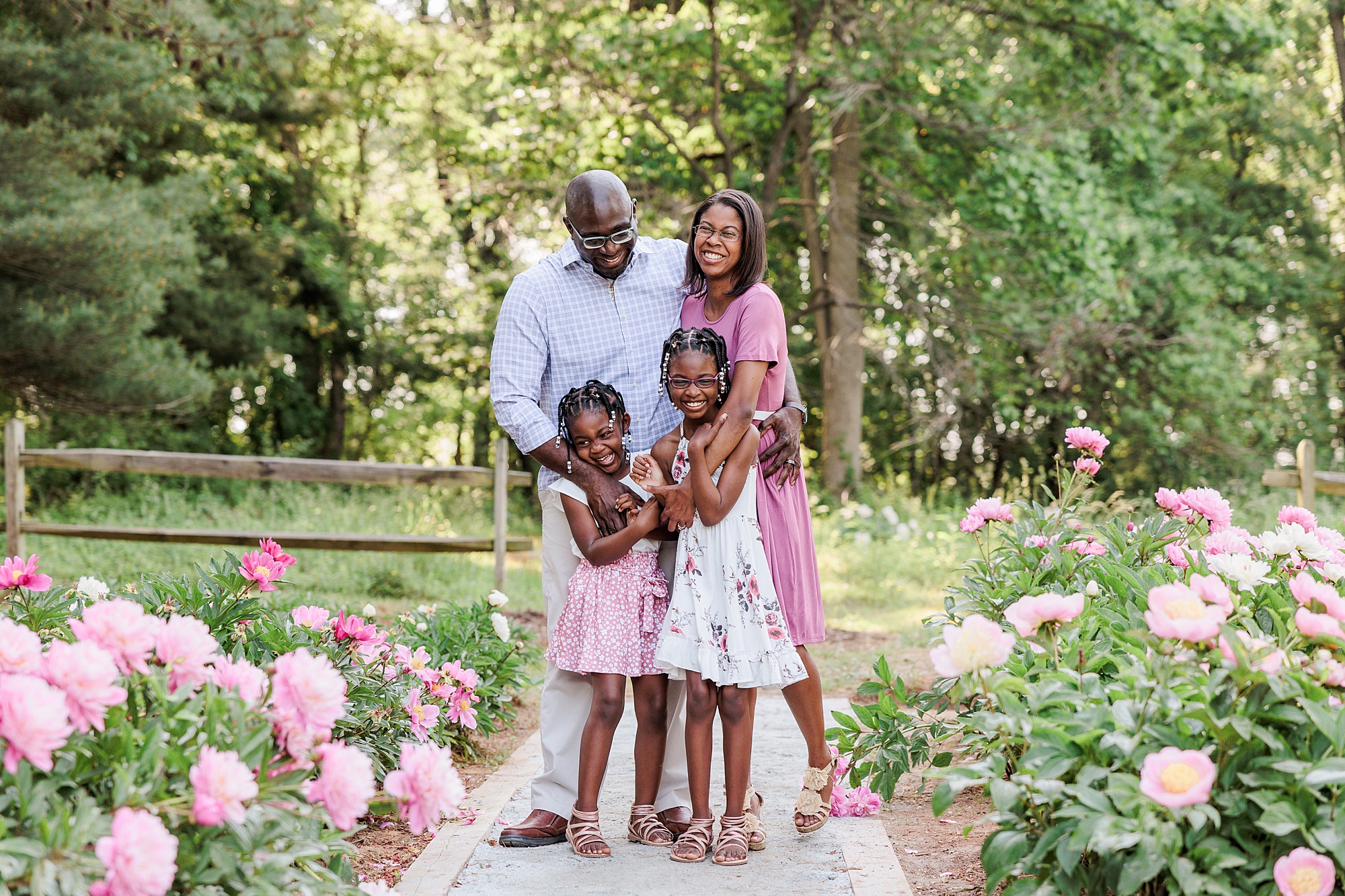 parents stand with daughters in pink and white dresses between rows of peonies