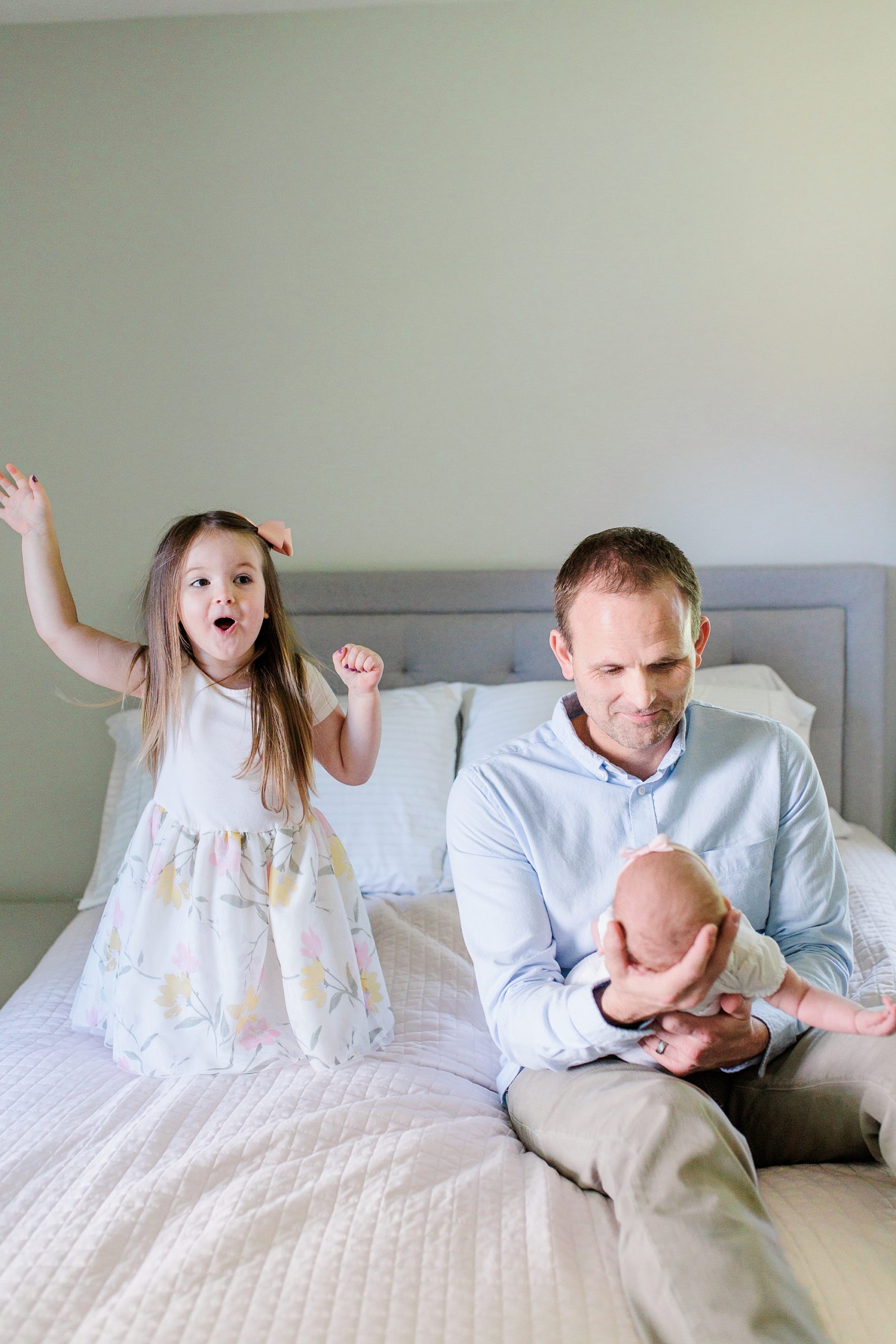 dad holds baby while big sister jumps on bed next to them