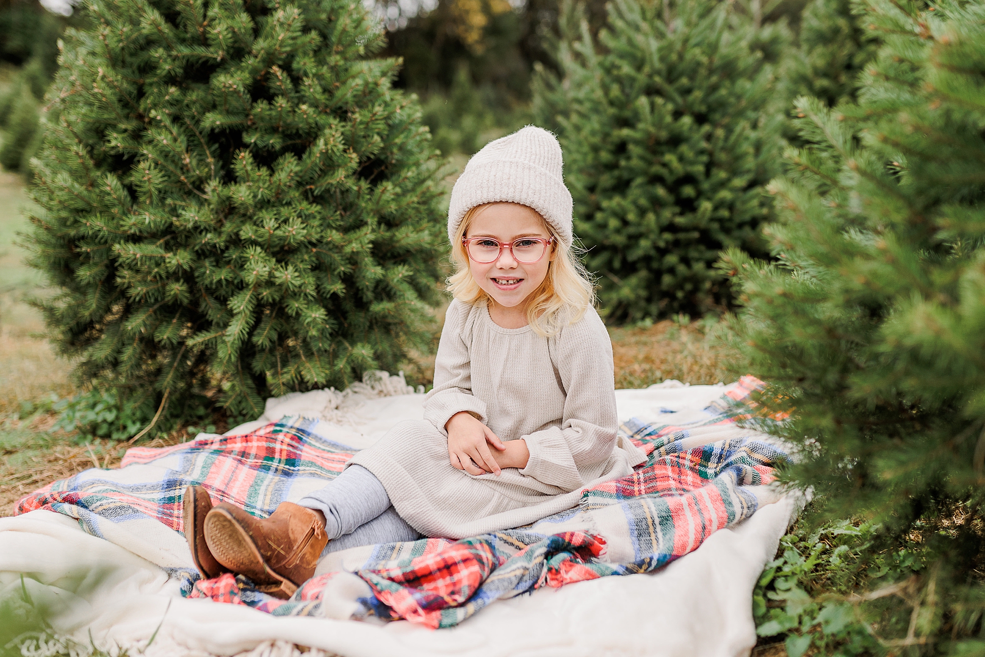 girl in ivory beanie sits on blanket between Christmas trees
