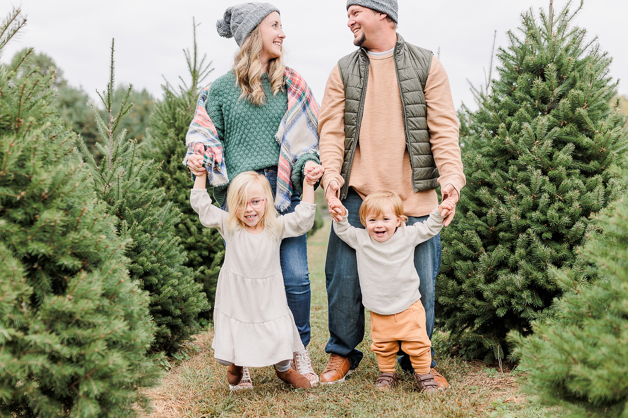 couple holds kids' hands while looking at each other during Christmas tree farm portraits