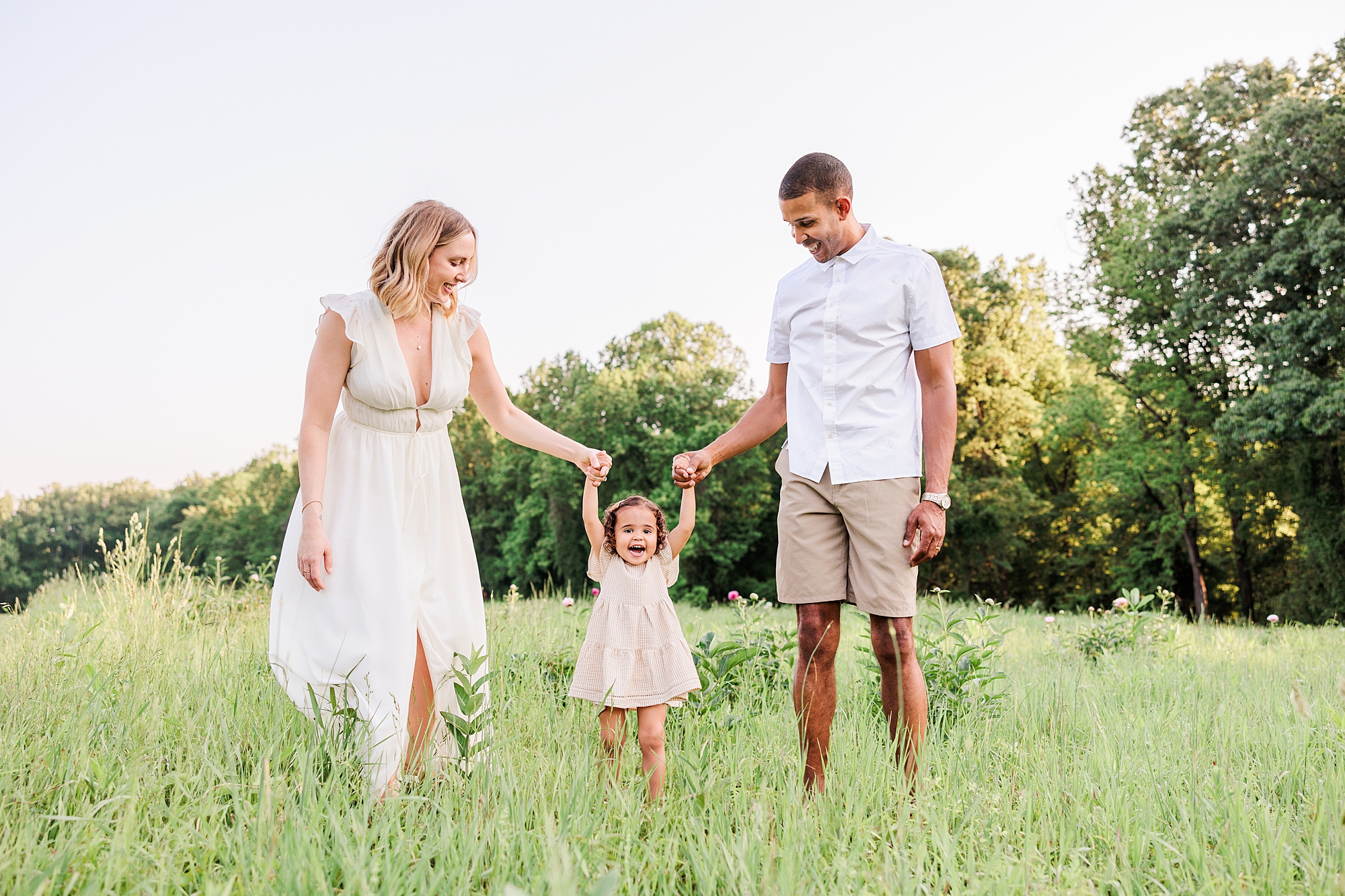 mom in ivory dress holds hands with toddler in cream dress in field with dad on other side 