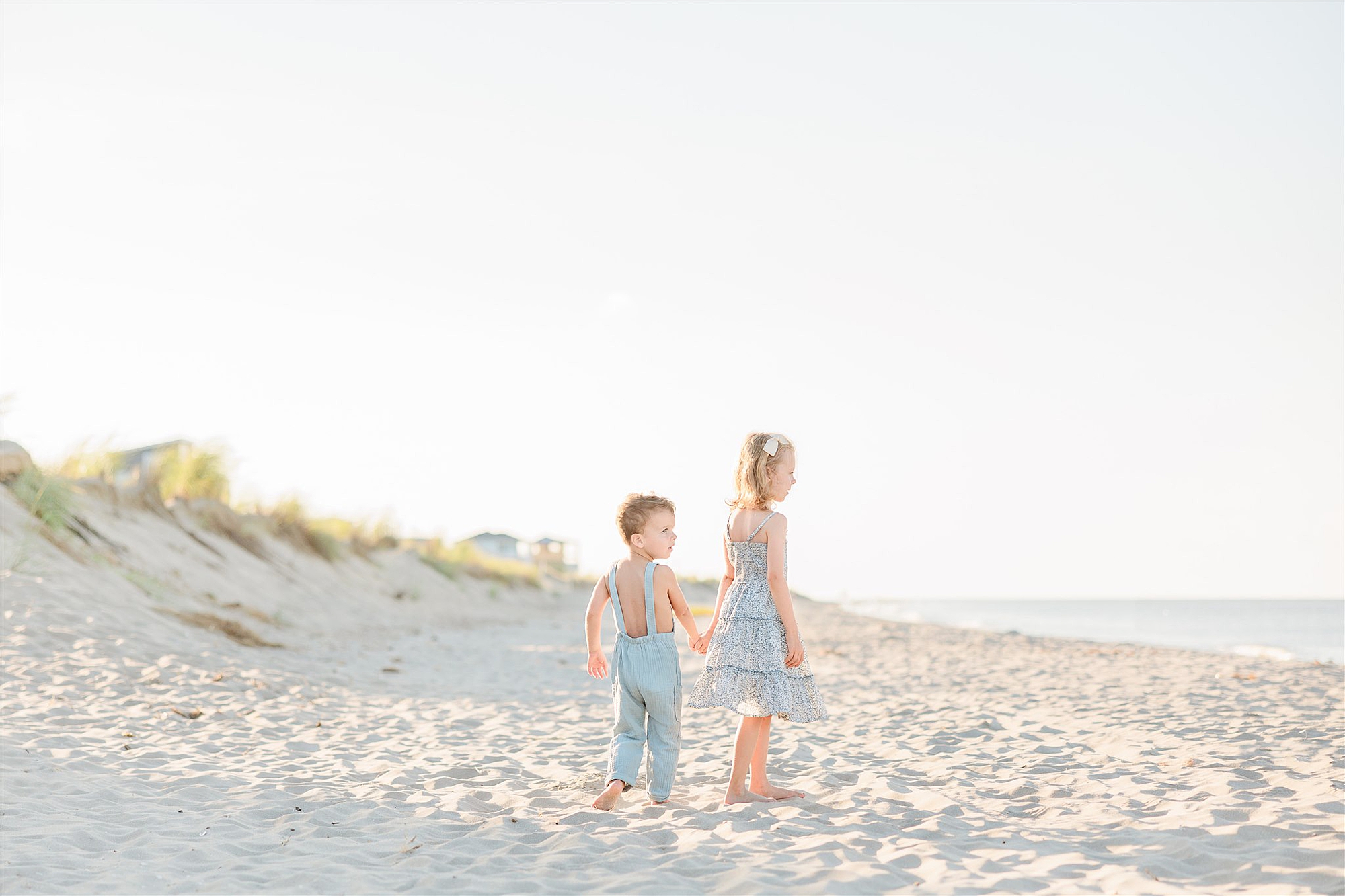 blonde girl leads boy in blue overalls across sand on beach 
