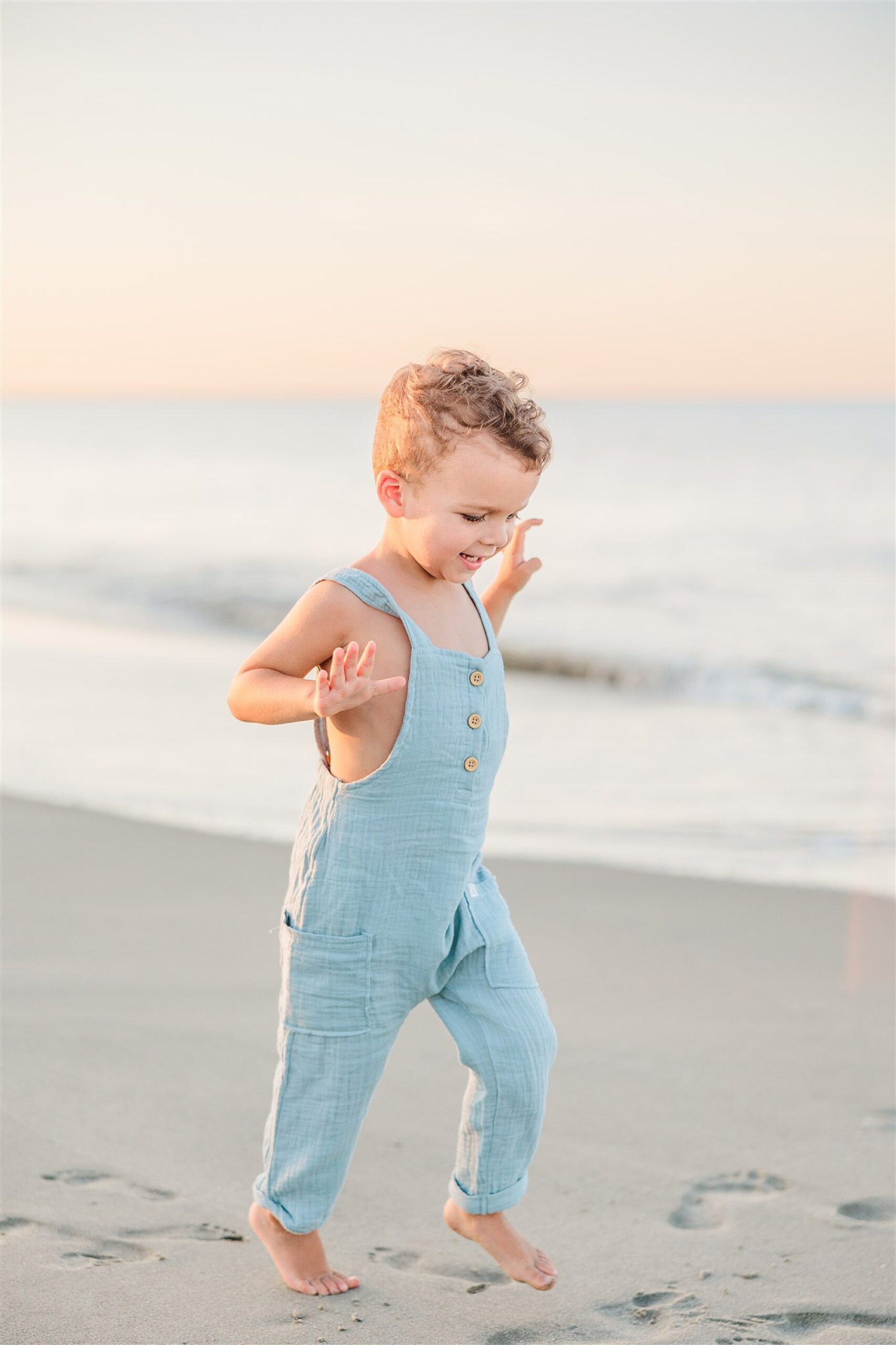toddler boy runs on sand in blue overalls 