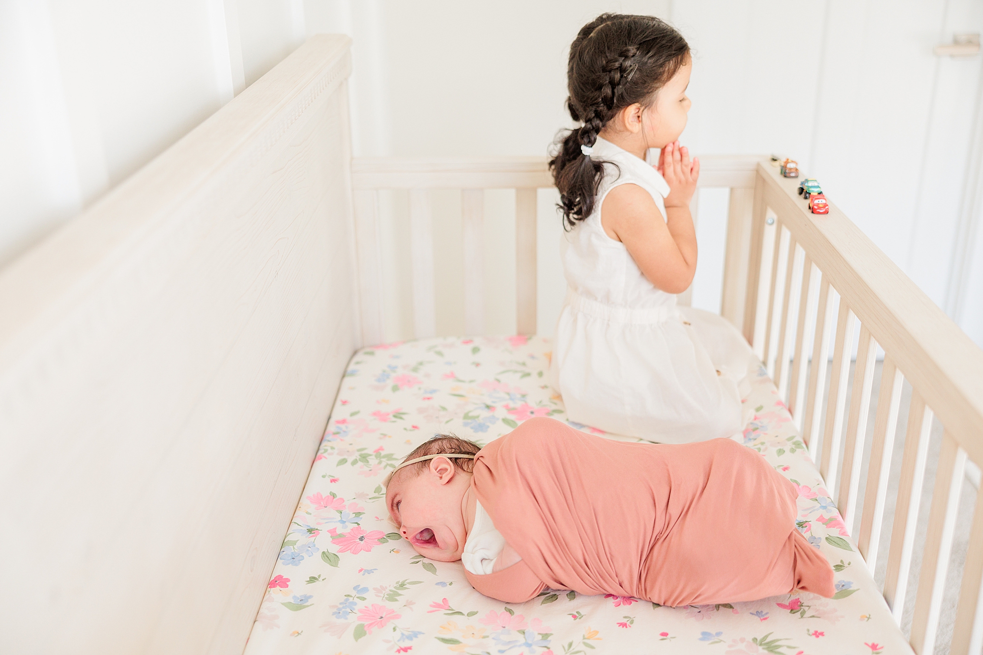 big sister leans on edge of railing in crib while baby sisters sleeps next to him 