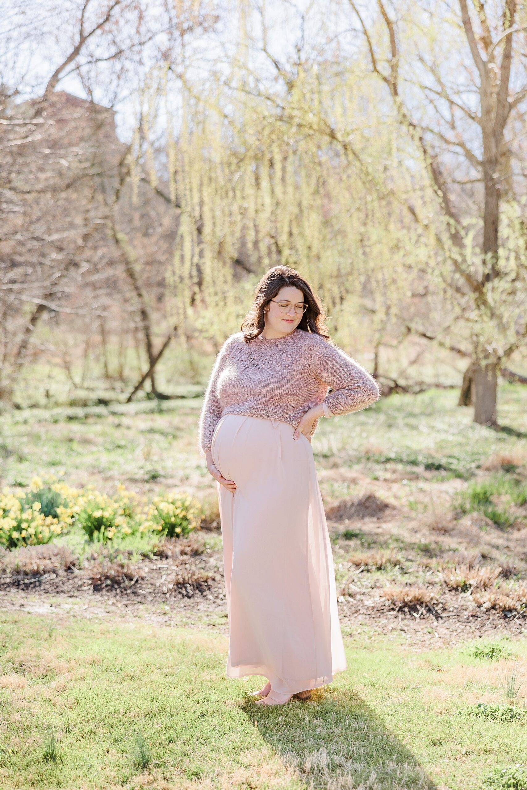 expecting mom stands in pink maternity gown with knit sweater on top at Lake Kittamaqundi
