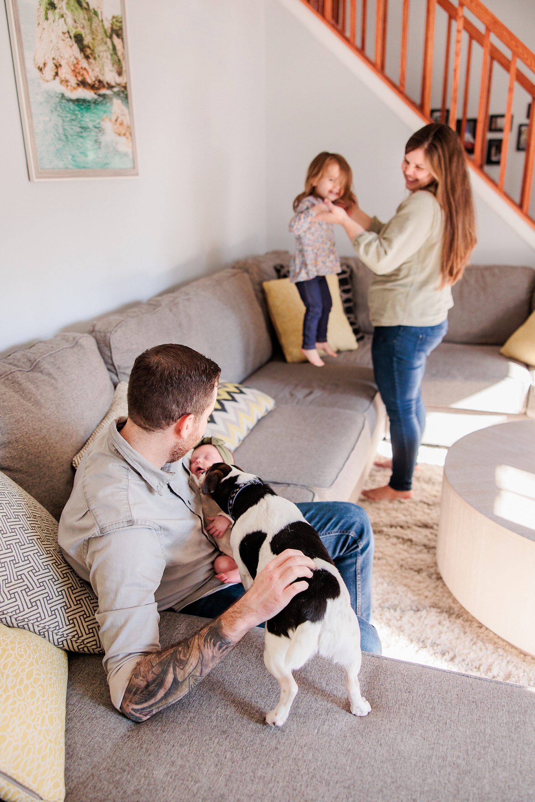 parents play with toddler and dog while holding newborn baby girl on couch