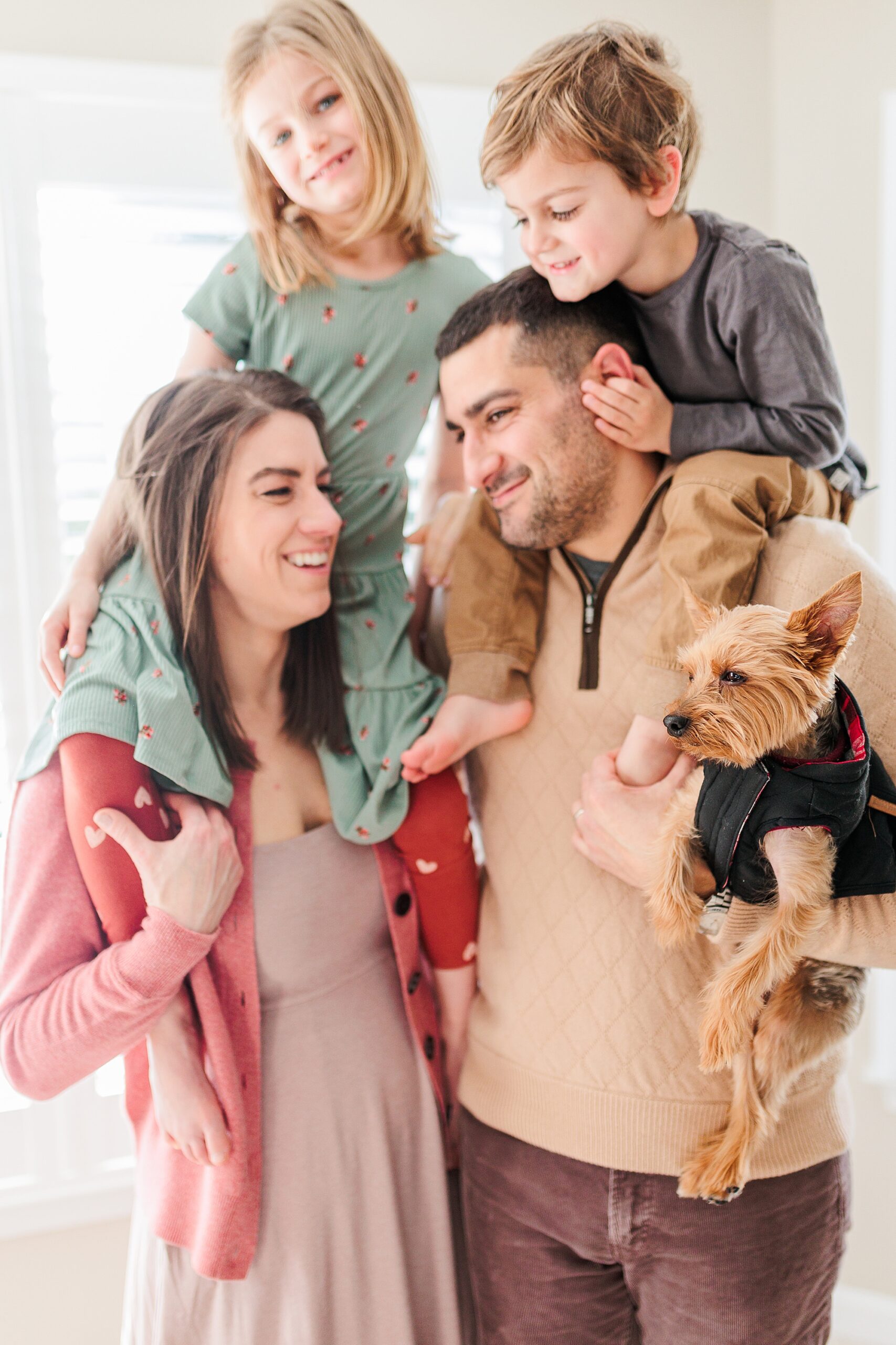 parents smile at each other with kids on their shoulders and holding small dog
