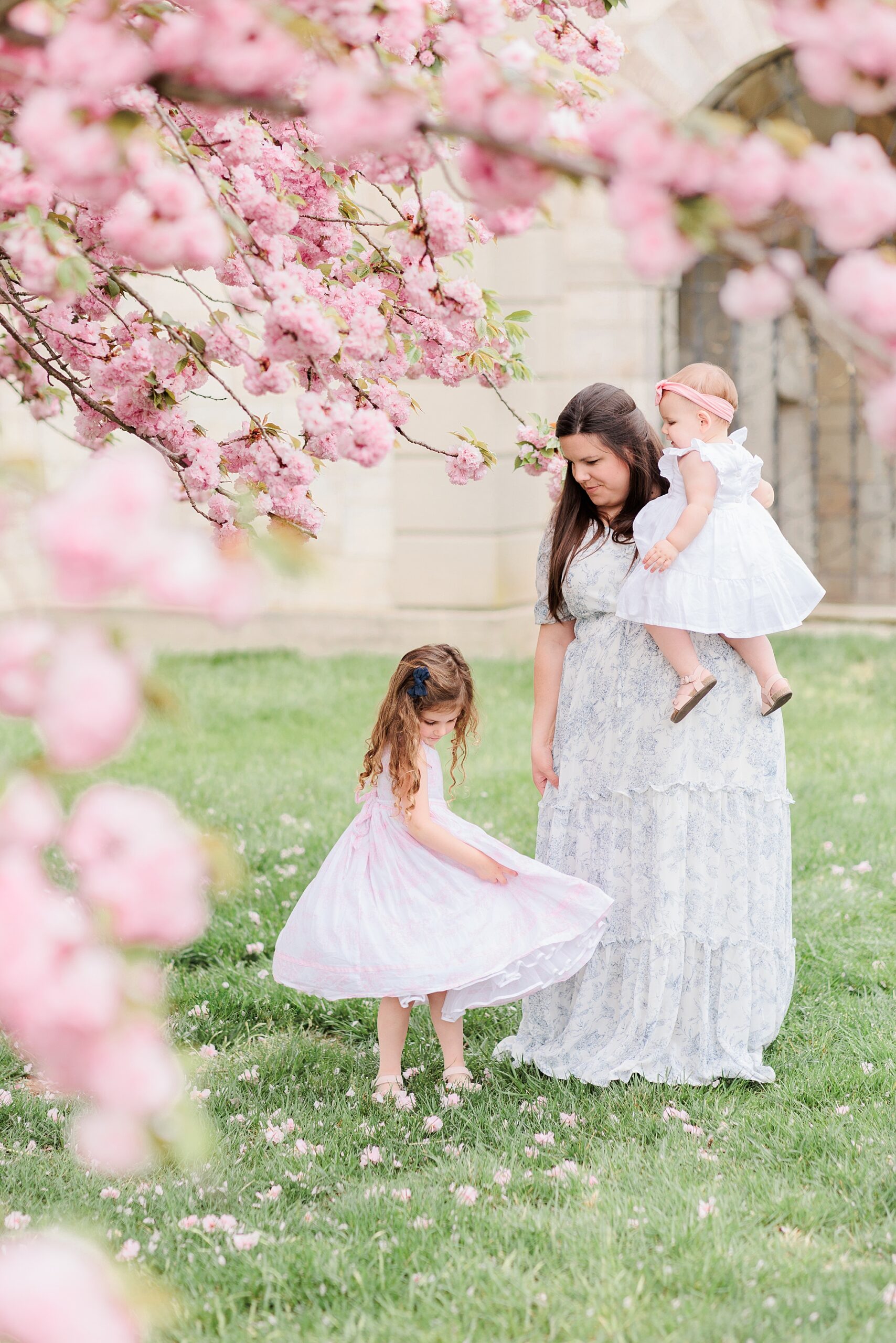 mother stands with two daughters under pink cherry blossom trees