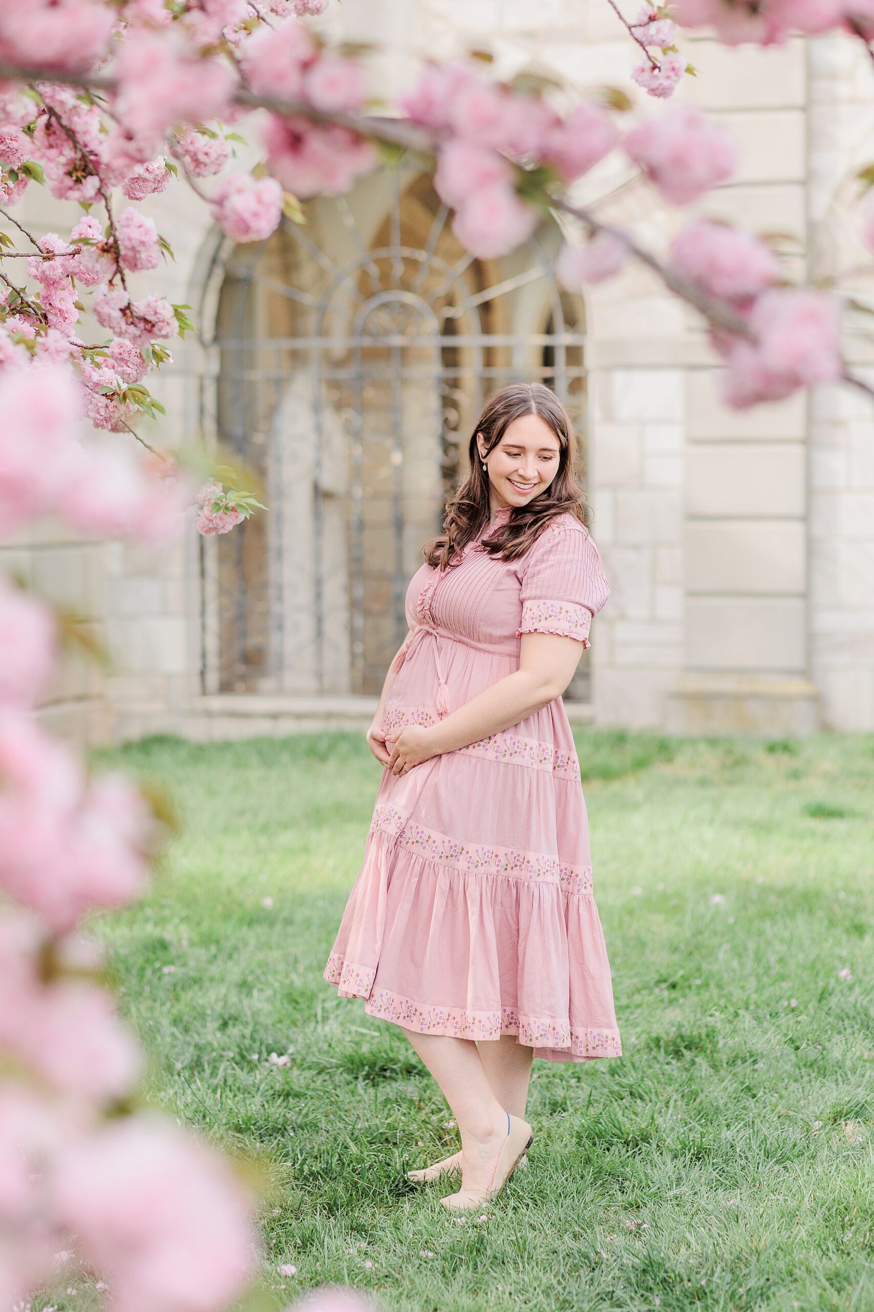 expecting mom in pink dress holds baby bump under spring blossoms 
