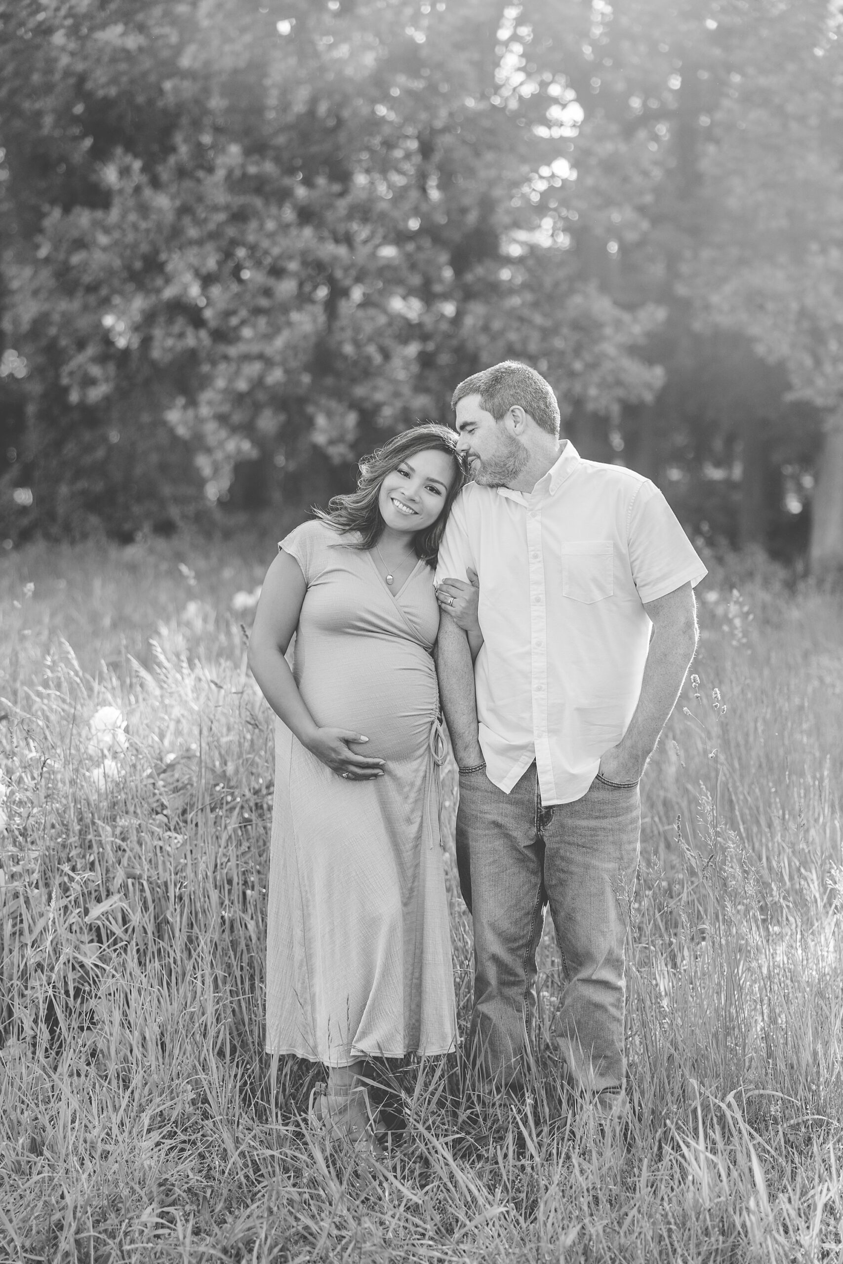 woman holds baby bump while leaning on husband's shoulder standing in field at Seneca Creek Park