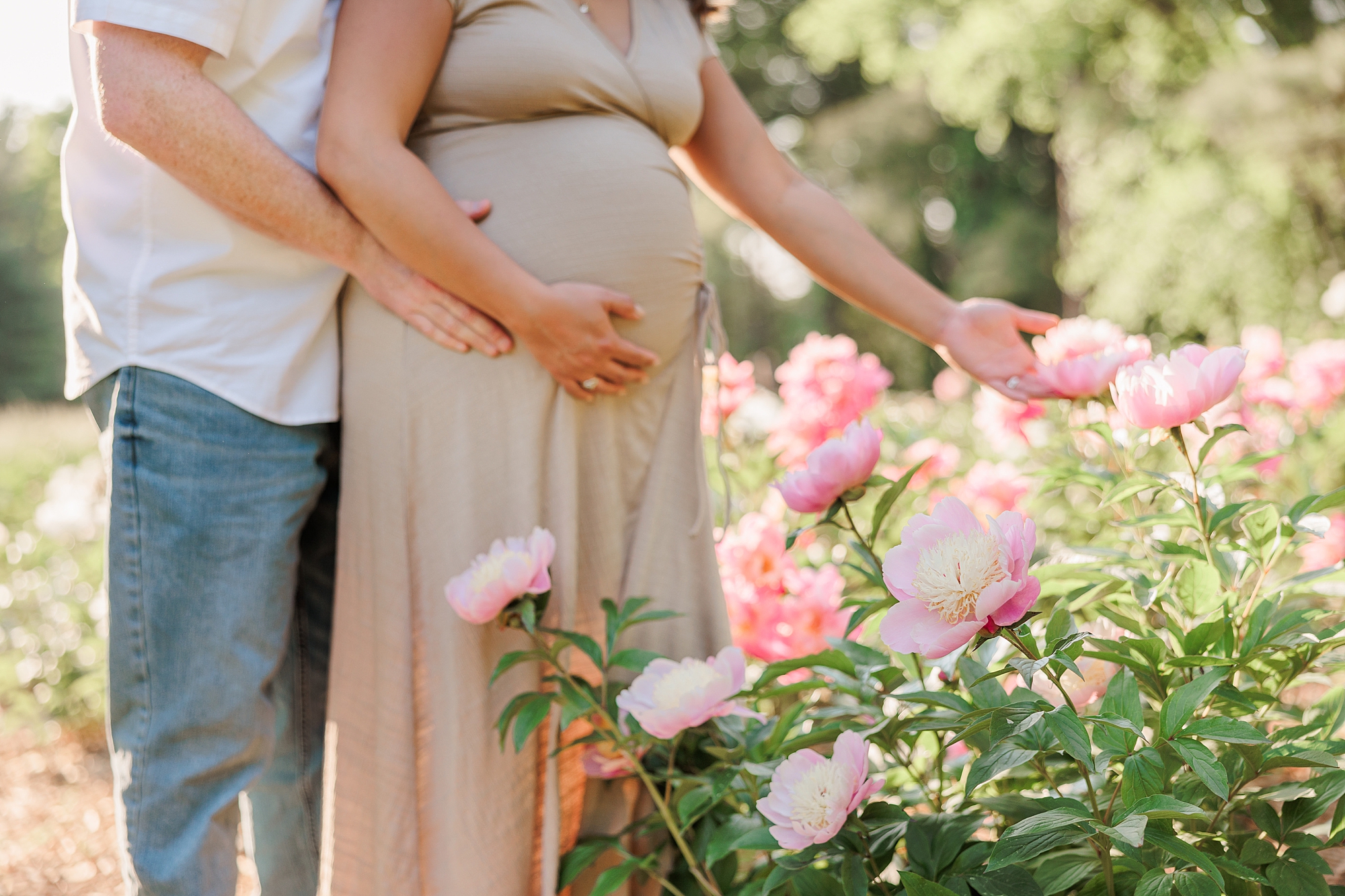 couple looks at pink peonies at Seneca Creek Park while holding onto baby bump