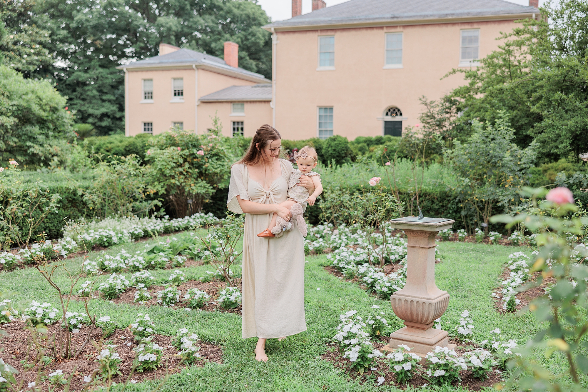mom walks through garden during Tudor Place family photos with toddler daughter on hip