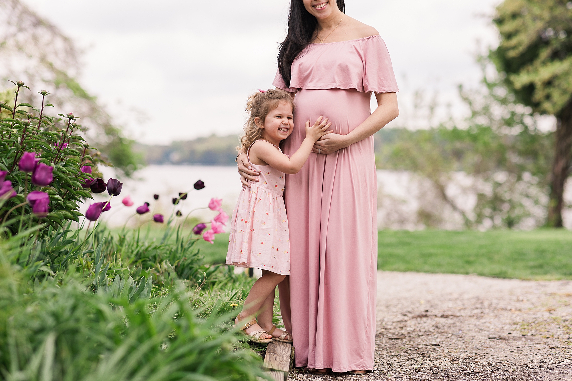 mom in pink gown holds baby bump with oldest daughter hugging her