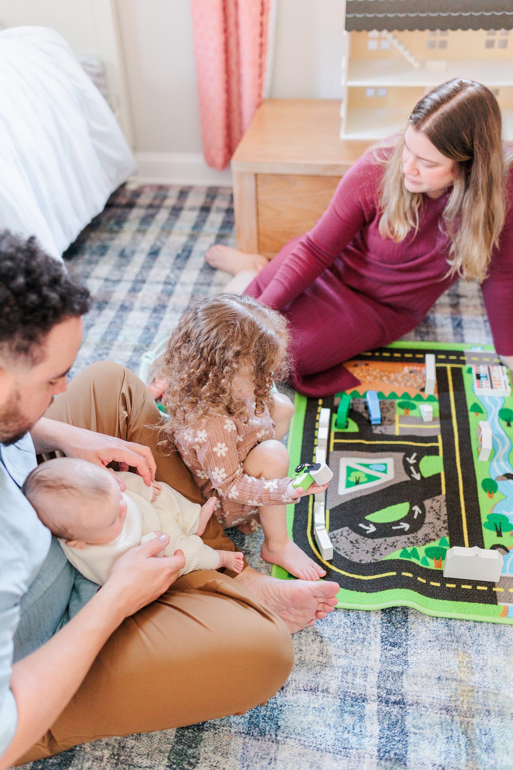 parents play with race track with toddler daughter while baby sits in dad's lap