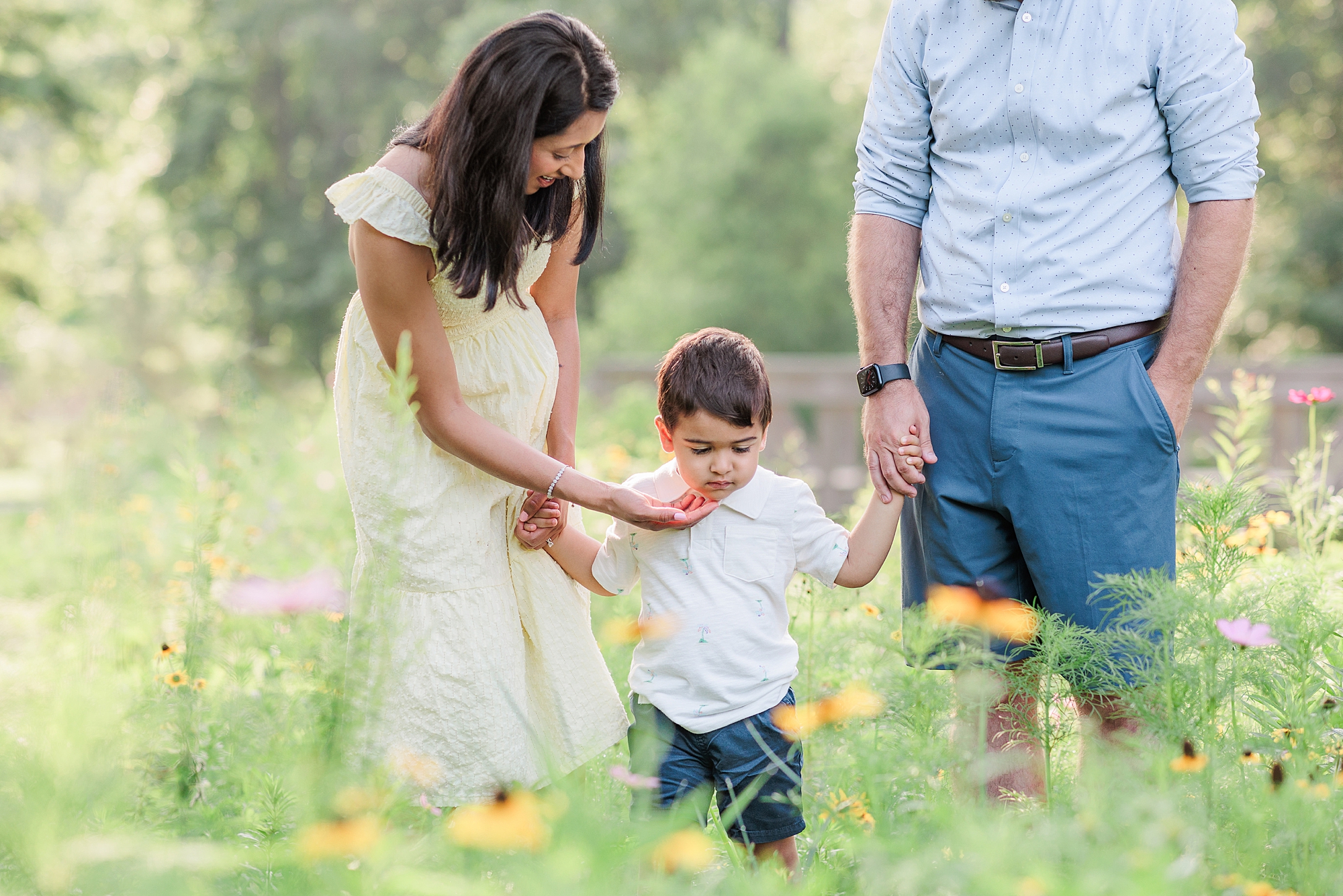 parents hold son's hand walking through field of wildflowers 