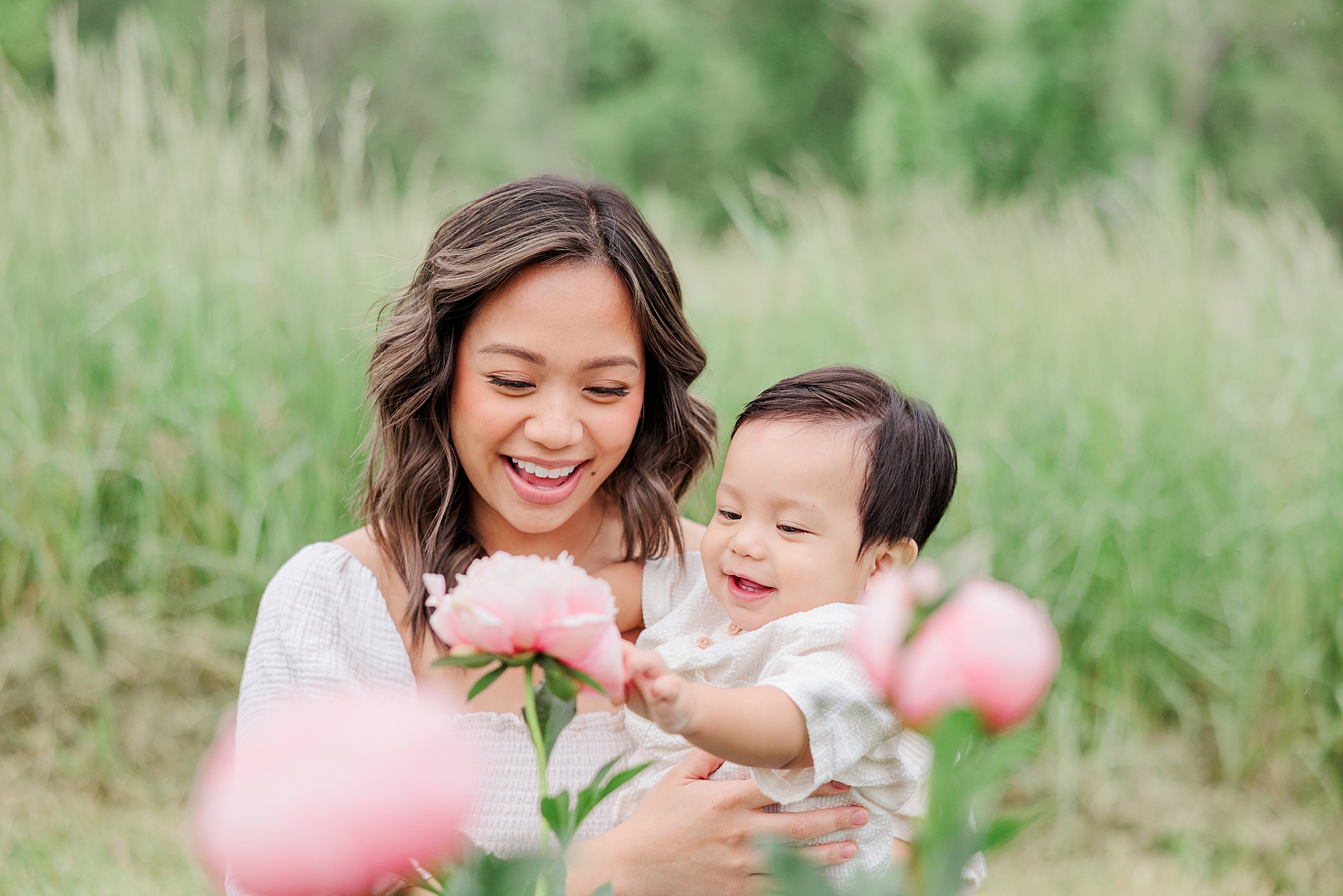 mom laughs holding toddler son in field of pink peonies 