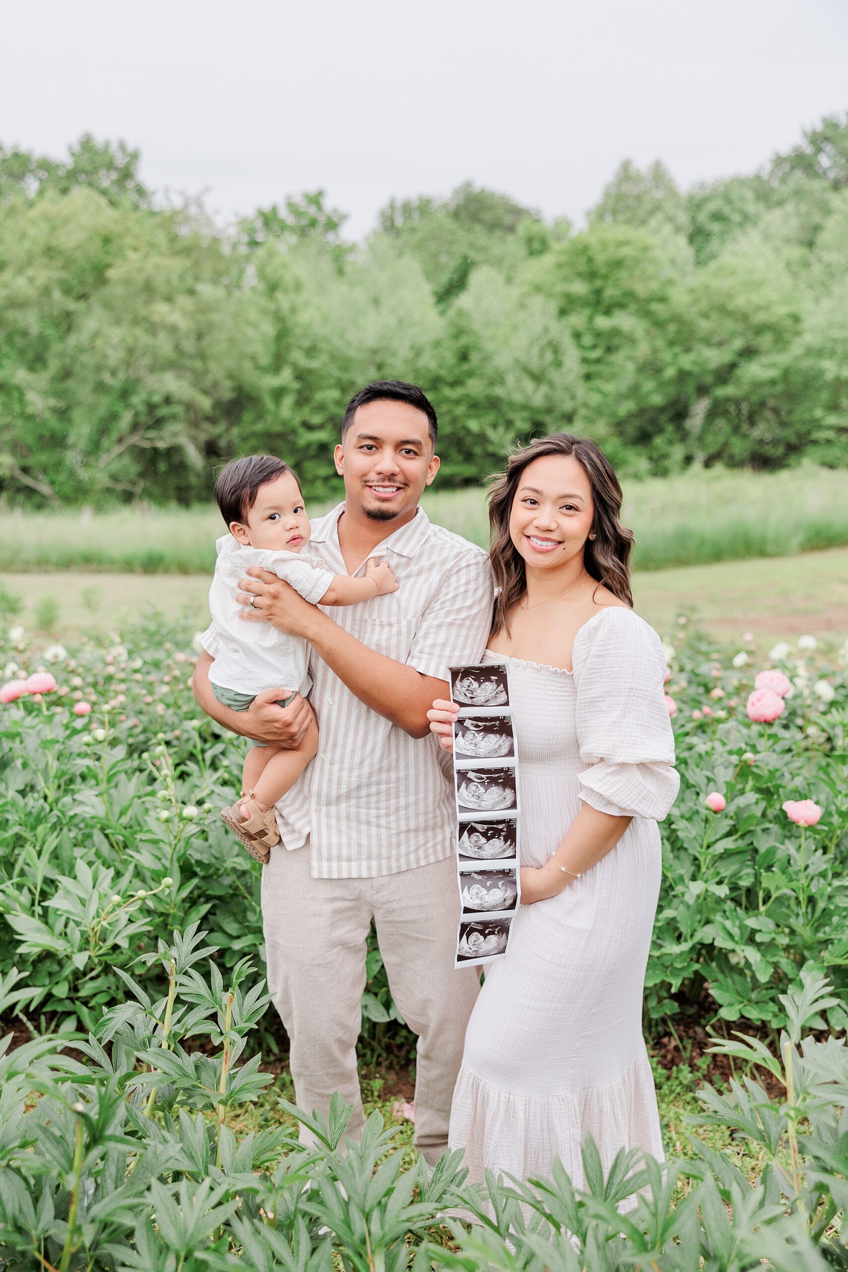 parents stand holding toddler while mom shows off sonogram