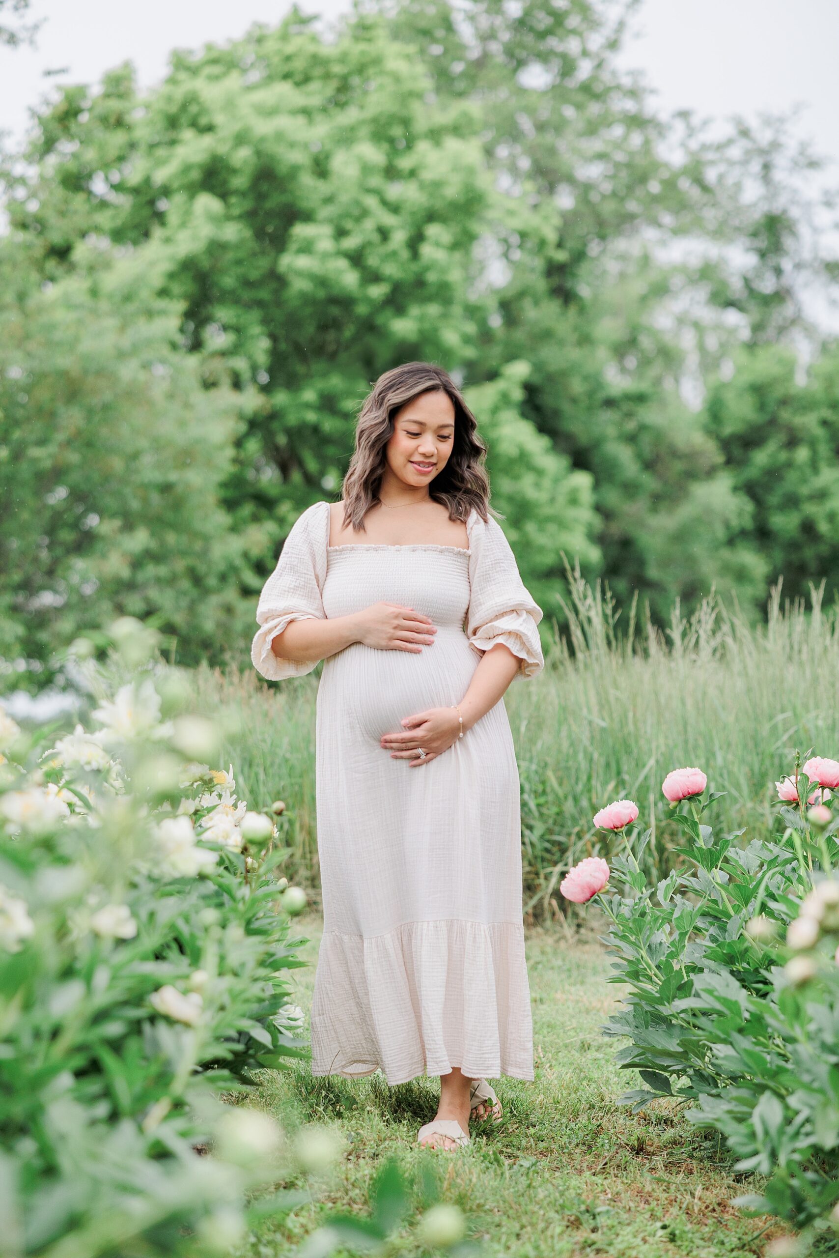 woman in white dress stands between peonies during pregnancy announcement at Castlebridge Farm