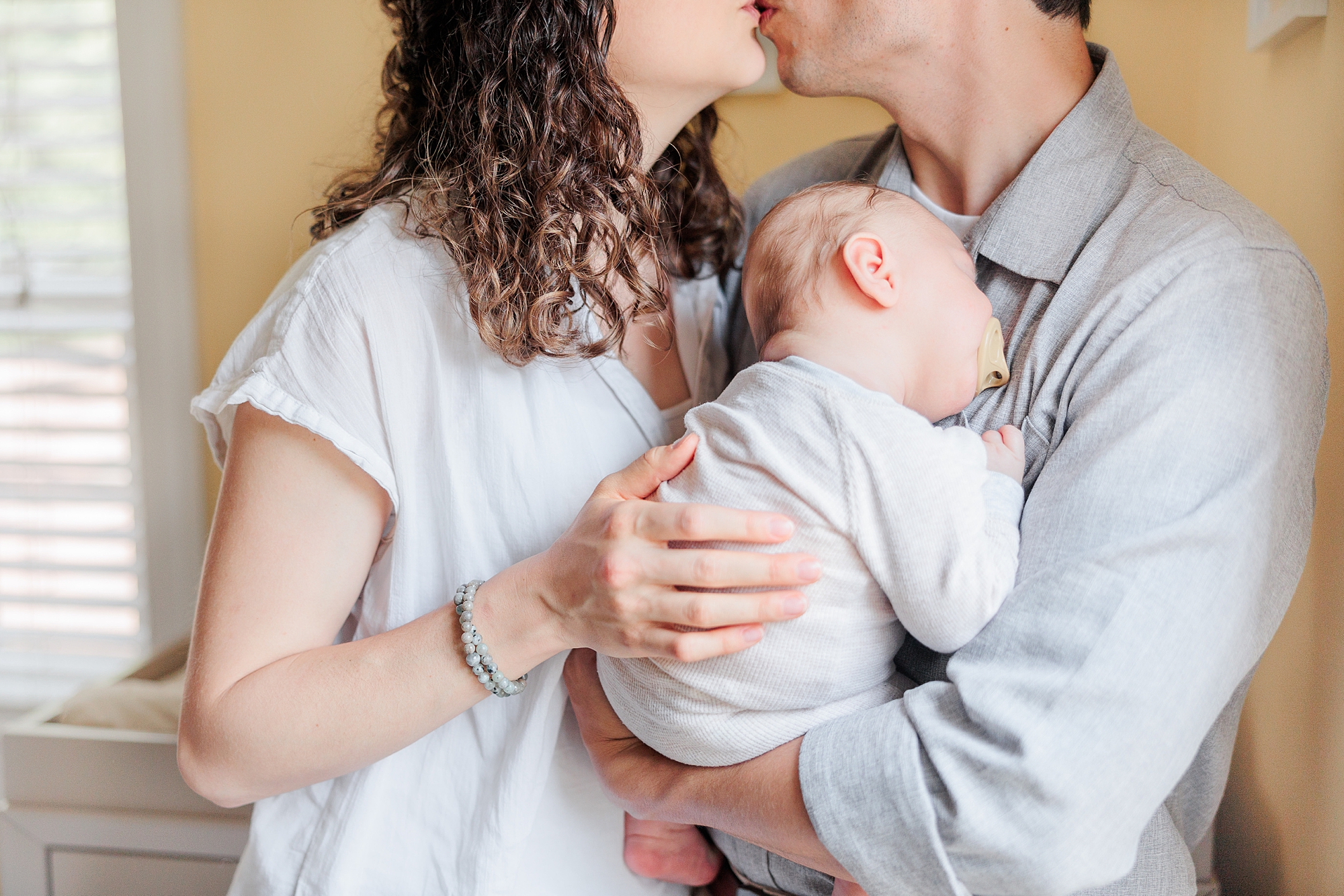 newlyweds kiss holding newborn son to chest during newborn lifestyle photography session at home