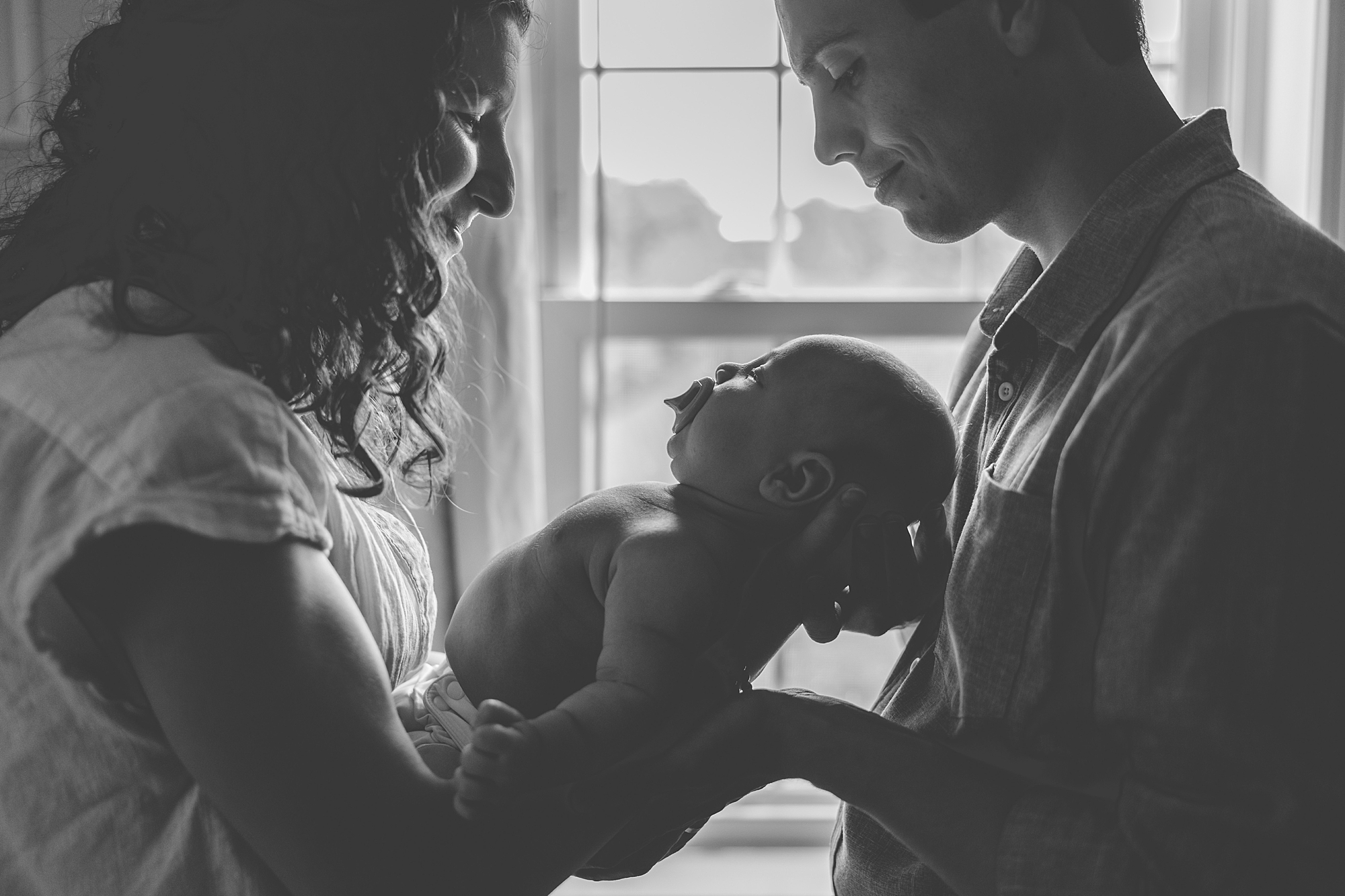 parents hold son between them in front of window 