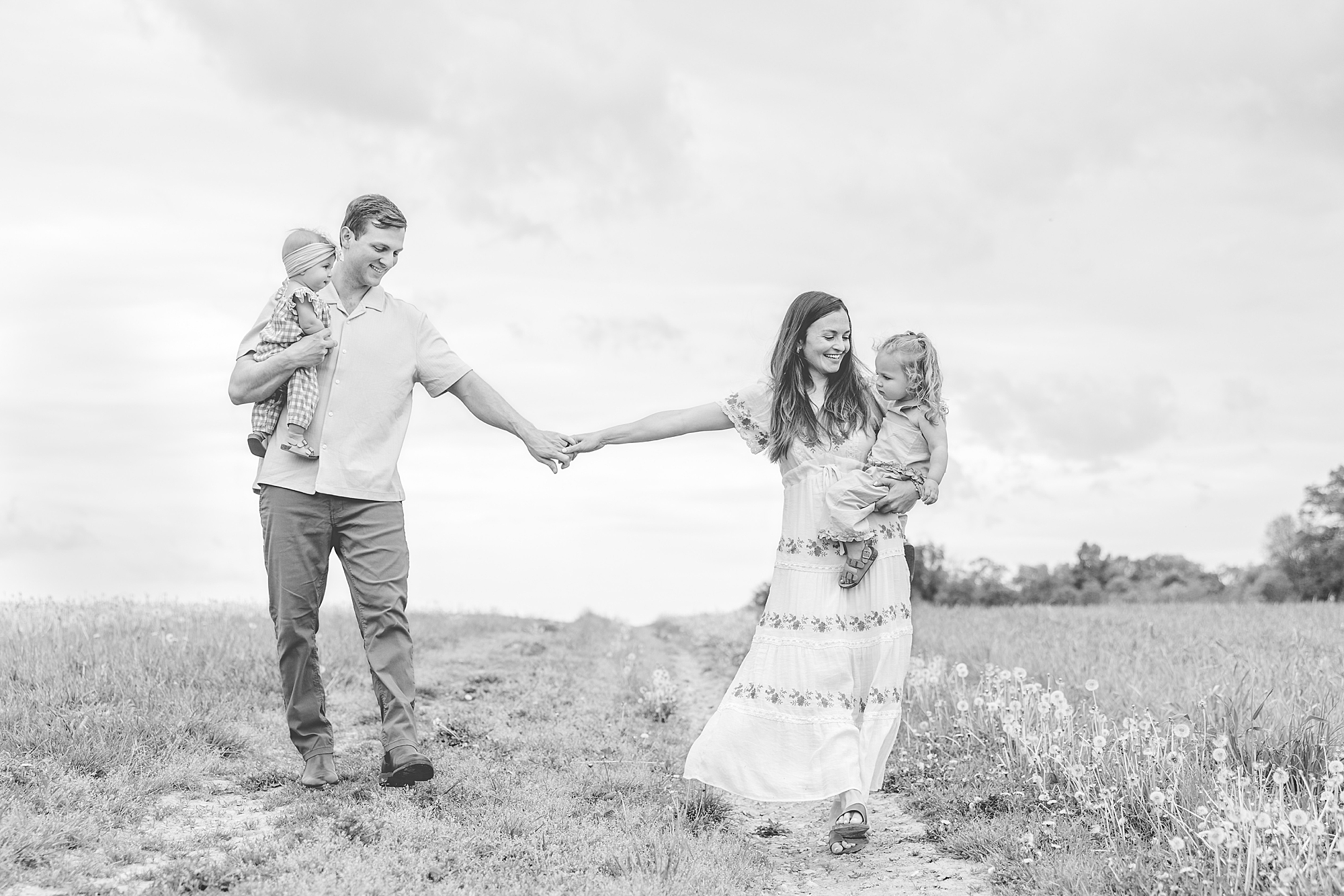 parents hold hands walking through field carrying two daughters 