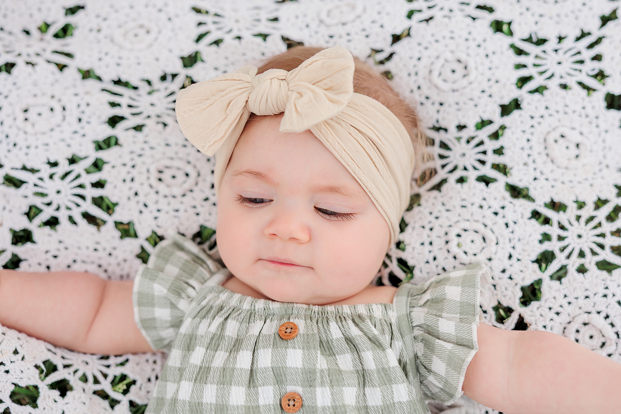 baby girl in tan bow lays on crocheted white blanket 