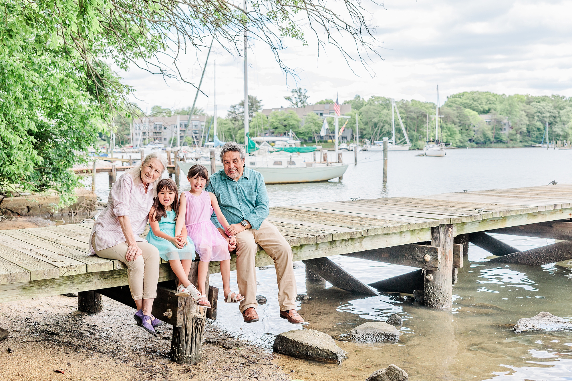 grandparents sit on dock with granddaughters during extended family session with Annapolis Maryland family photographer